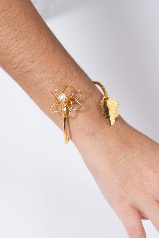 Pulsera dorada abierta con detalle de flor metálica y adorno lateral tipo hoja.
